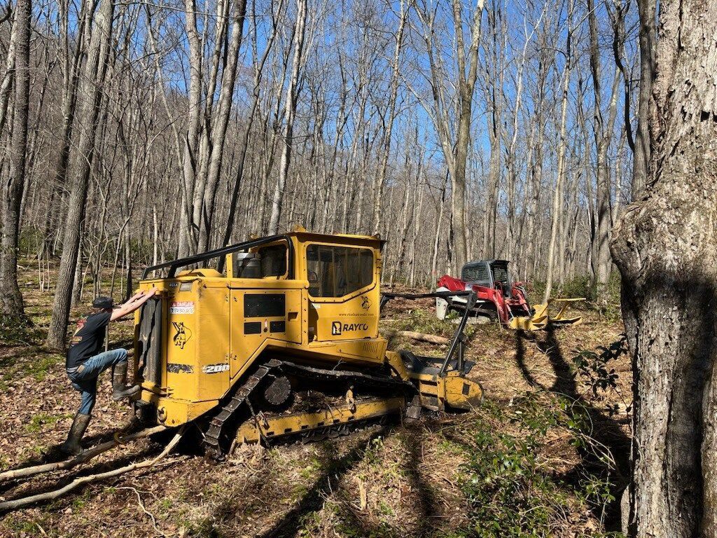 Yellow bulldozer pulling a red machine through a wooded area. Person in dark clothing is working on the bulldozer.