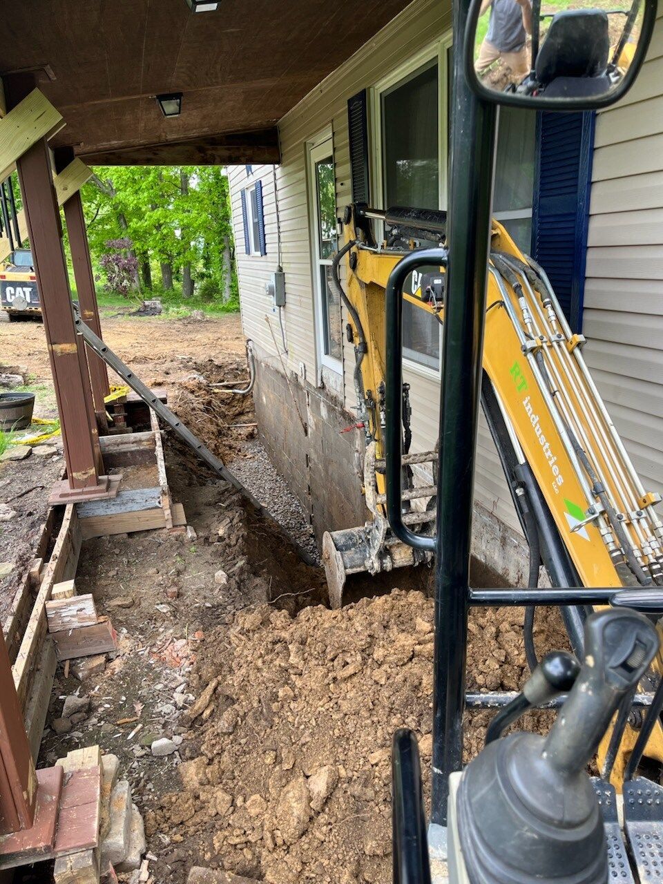 Mini excavator digging along the foundation of a house with brown siding and a porch.