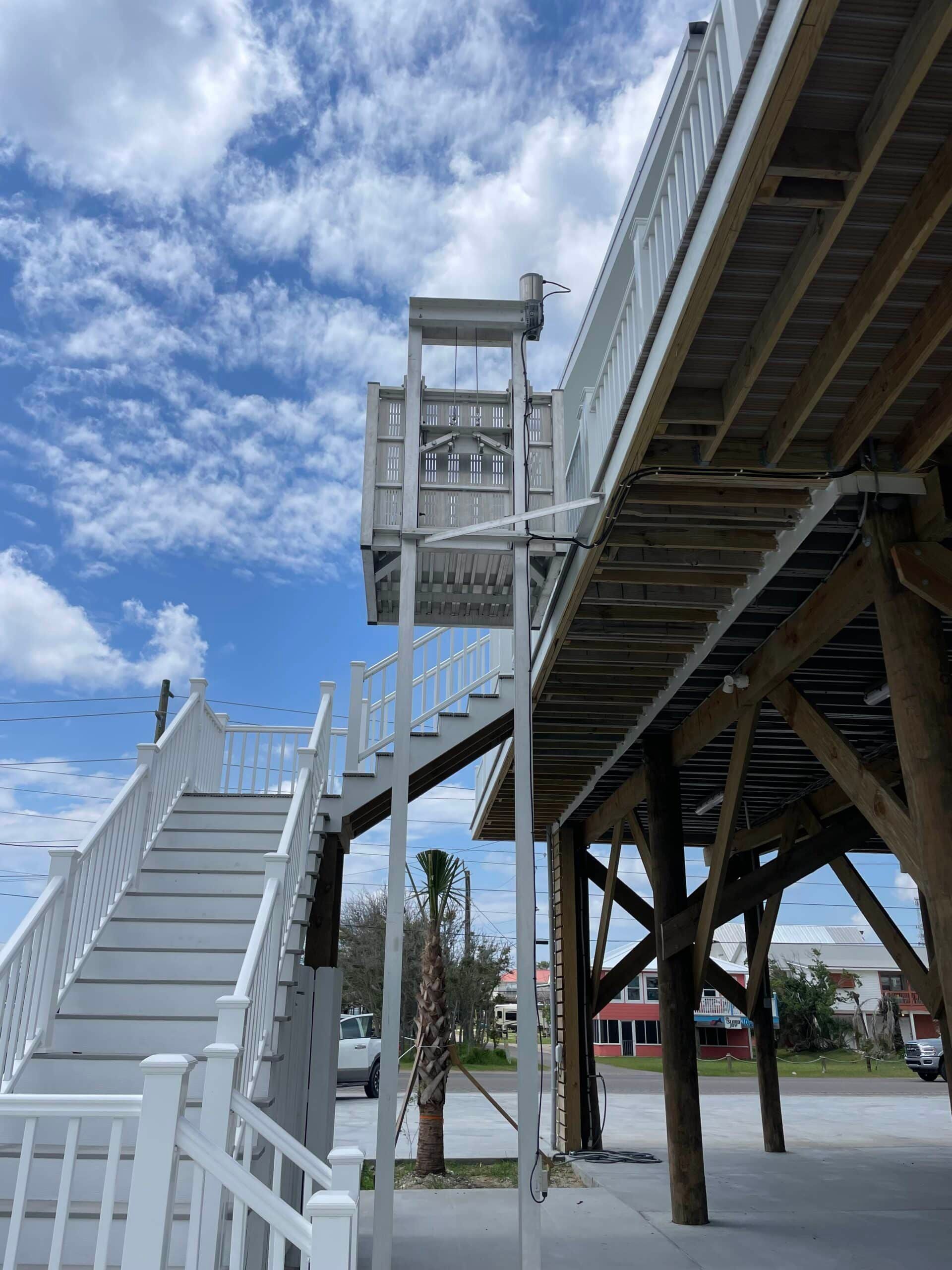 Elevated wooden pier with stairs and a elevator lift. White railings and supports against a blue sky.