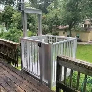 Outdoor elevator lift on a wooden deck. Aluminum lift with gated enclosure, attached to a house and overlooking a yard.