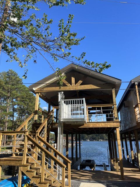 Wooden boat dock with a elevator lift, stairs, and a partial roof, overlooking a lake under a blue sky.