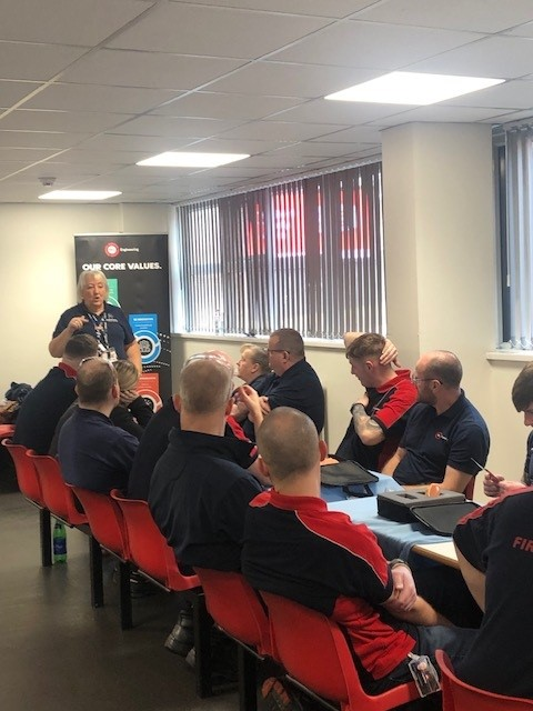 A group of people in red and blue shirts listen to a person speaking in a meeting room.