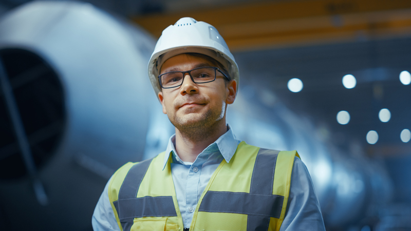 Man in safety vest and helmet, smiling in a factory setting, near large equipment.