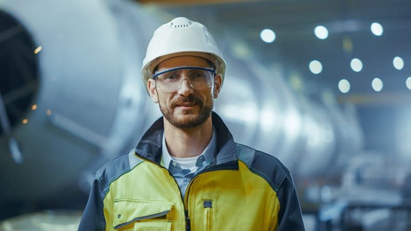 Man in hard hat and safety glasses smiles in a factory setting.