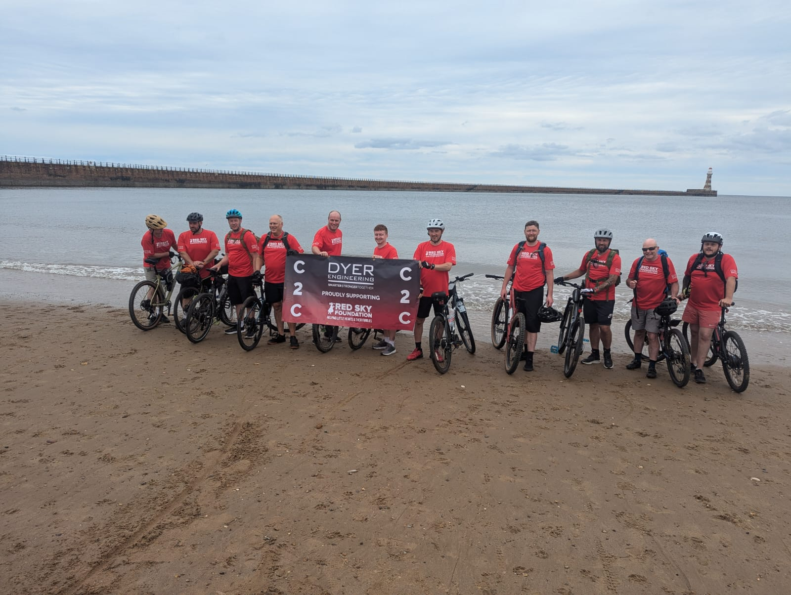 Group of cyclists in red shirts standing on a beach holding a banner near the water.
