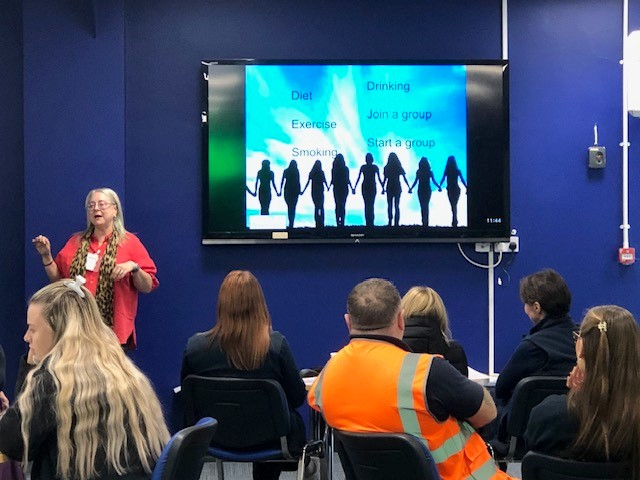 Woman presents to an audience in front of a screen with silhouetted figures holding hands, inside a room.