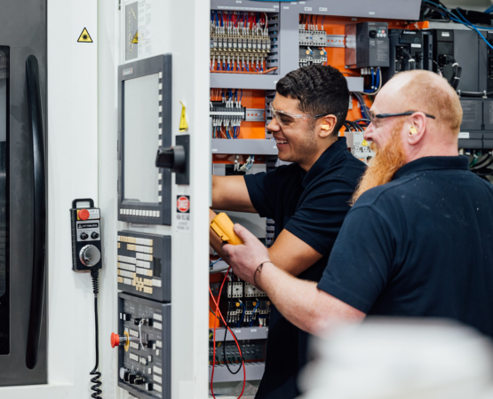 Two technicians in black shirts smiling as they work on electrical panel. One holds a multimeter.
