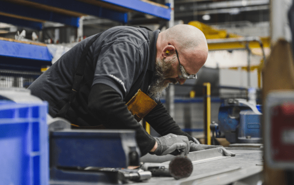 Man with beard and glasses working at a workbench in a factory; he's wearing gloves and safety glasses.