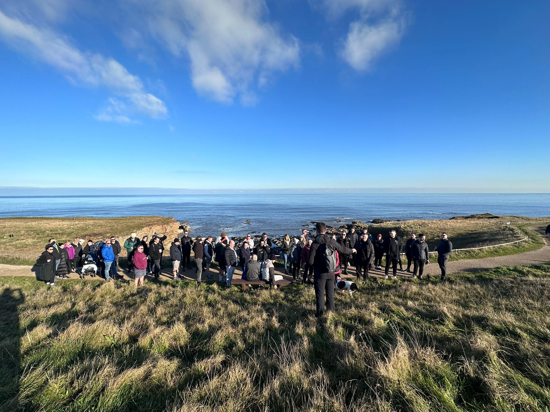 Group of people standing on a grassy cliff overlooking the ocean, raising their arms on a sunny day.