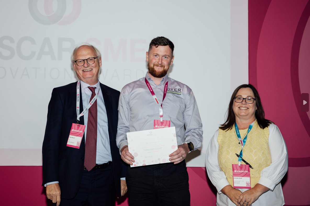 Three people on a stage: a man receiving an award from two people, all wearing lanyards, against a pink backdrop.