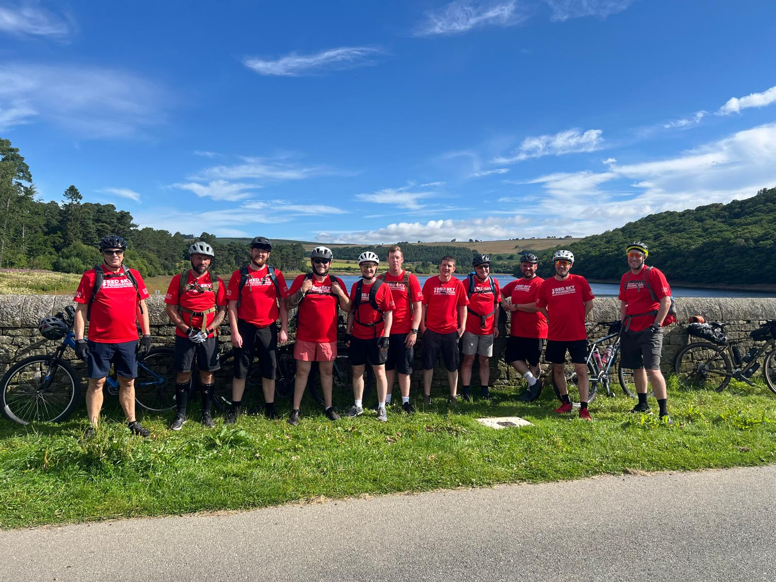 Cyclists in red shirts pose by a stone wall, lake, and green hills under a blue sky.