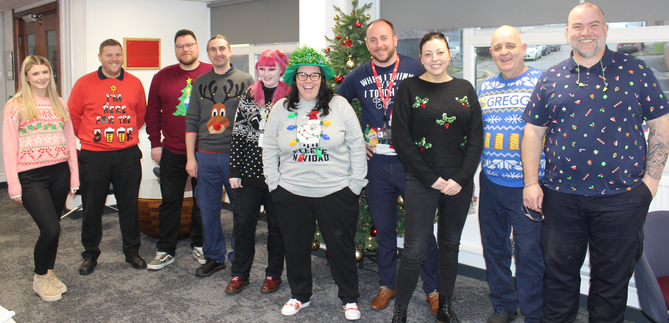 A group of people in Christmas sweaters stand in an office setting near a Christmas tree.