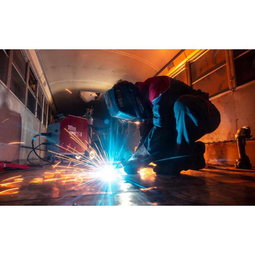 Welder inside a bus, sparks flying. Person in a helmet, welding, squatting down. Blue and orange lights.