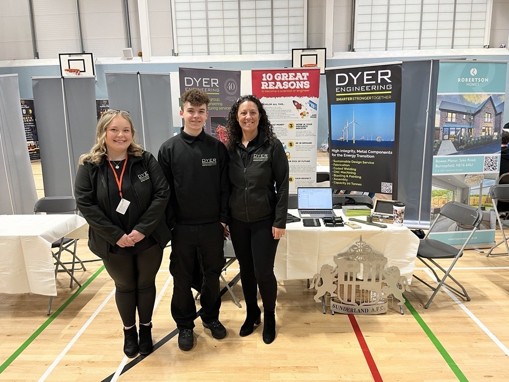 Three people at a trade show booth with company banners. Two women and a man stand in front of a table.