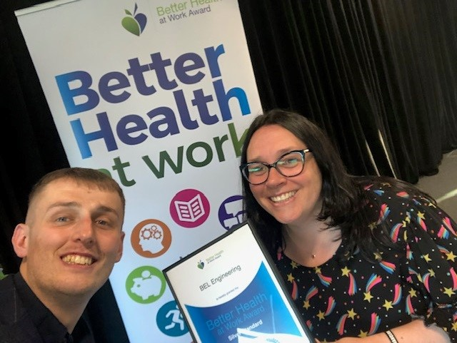 Two people smiling, holding a Better Health at Work award in front of a banner.