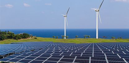 Solar panels and wind turbines on a grassy hillside overlooking the ocean under a blue sky.