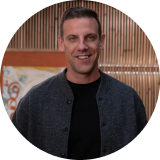 Smiling man in a dark jacket and black shirt, in front of a wooden wall and abstract art.