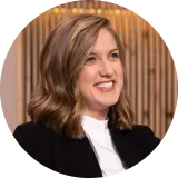 Woman with brown wavy hair smiles, wearing a black blazer and white shirt, in a warm-toned room.