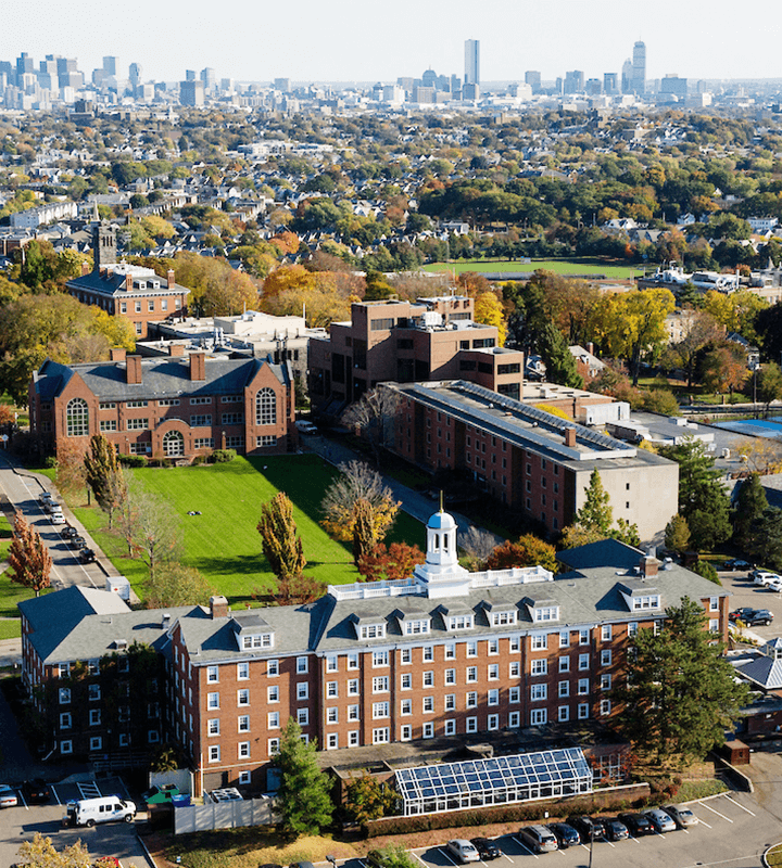 Aerial view of college campus with brick buildings, green lawns, and cityscape background.