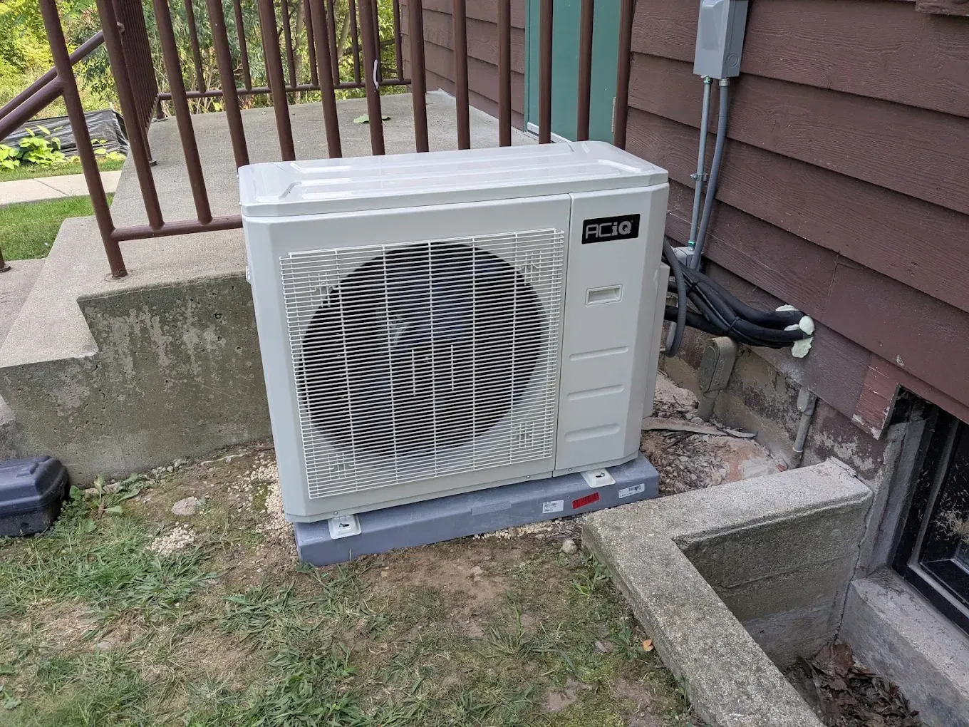 An outdoor heat pump unit on a gray base next to a brown house.