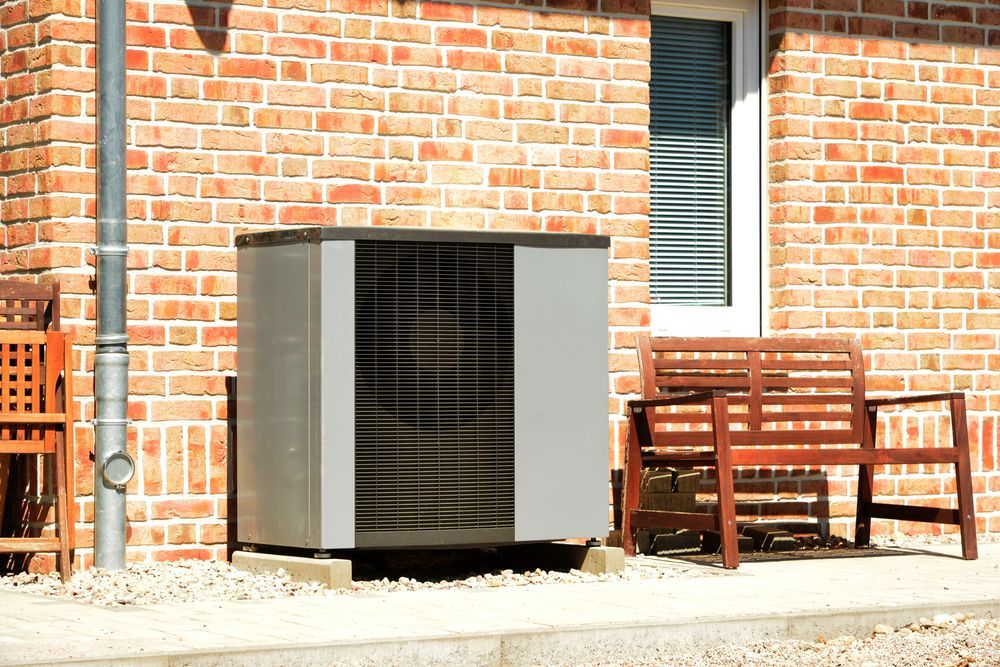 Heat pump unit outside a brick building, near a window and wooden bench.