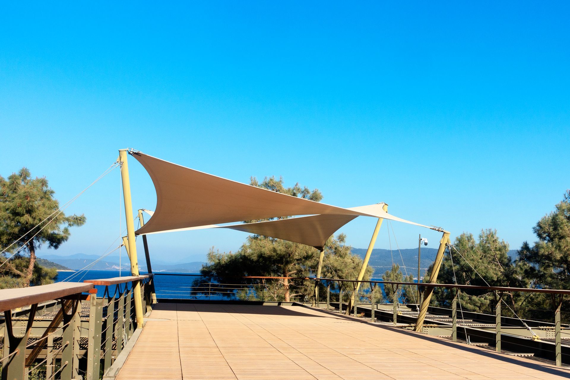 Sun shade sails on the tail end of a walking ramp with a blue sky, trees, and dark blue waters.