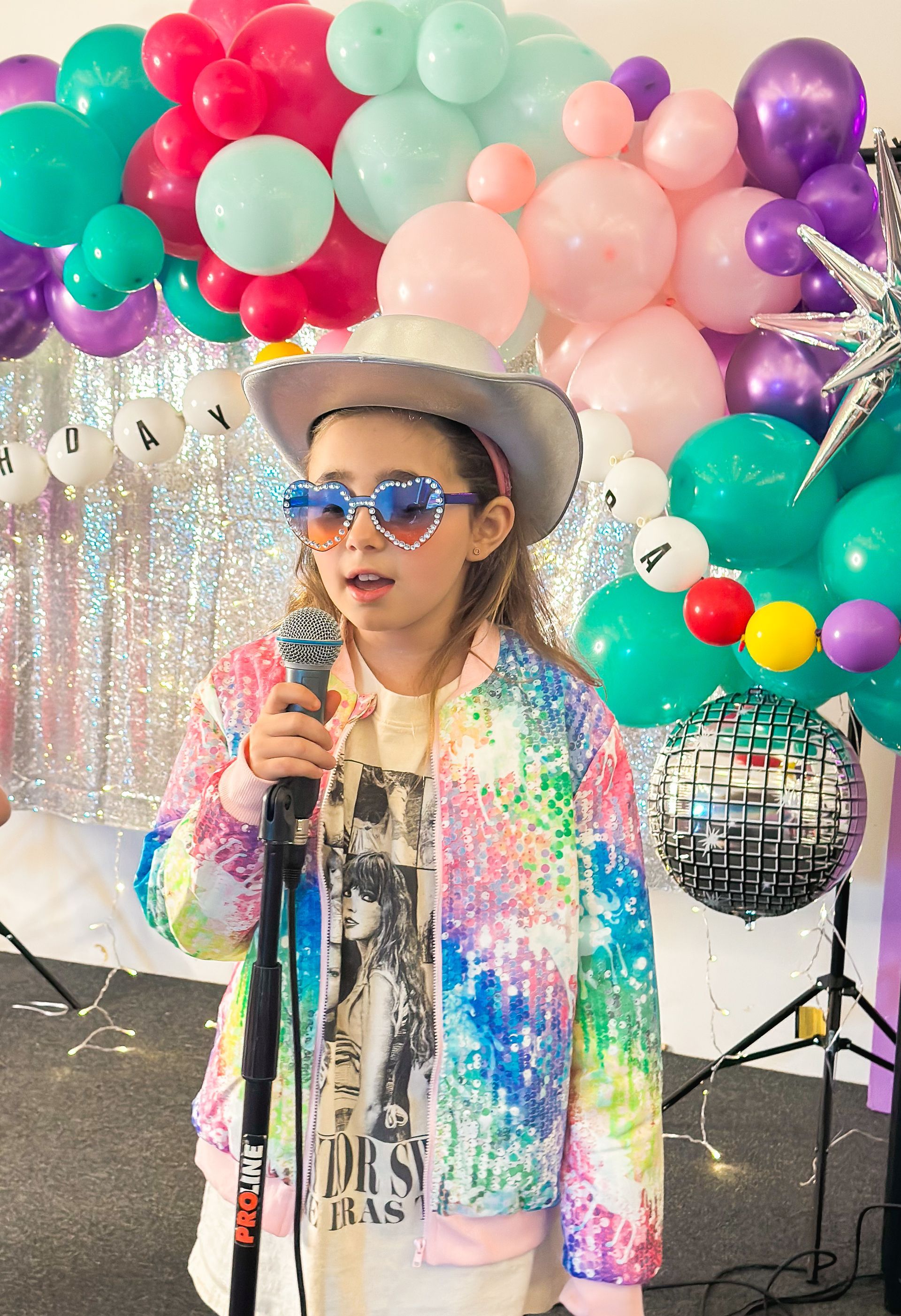 A little girl is singing into a microphone in front of balloons.