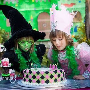 Two little girls dressed as witches are blowing out candles on a cake.