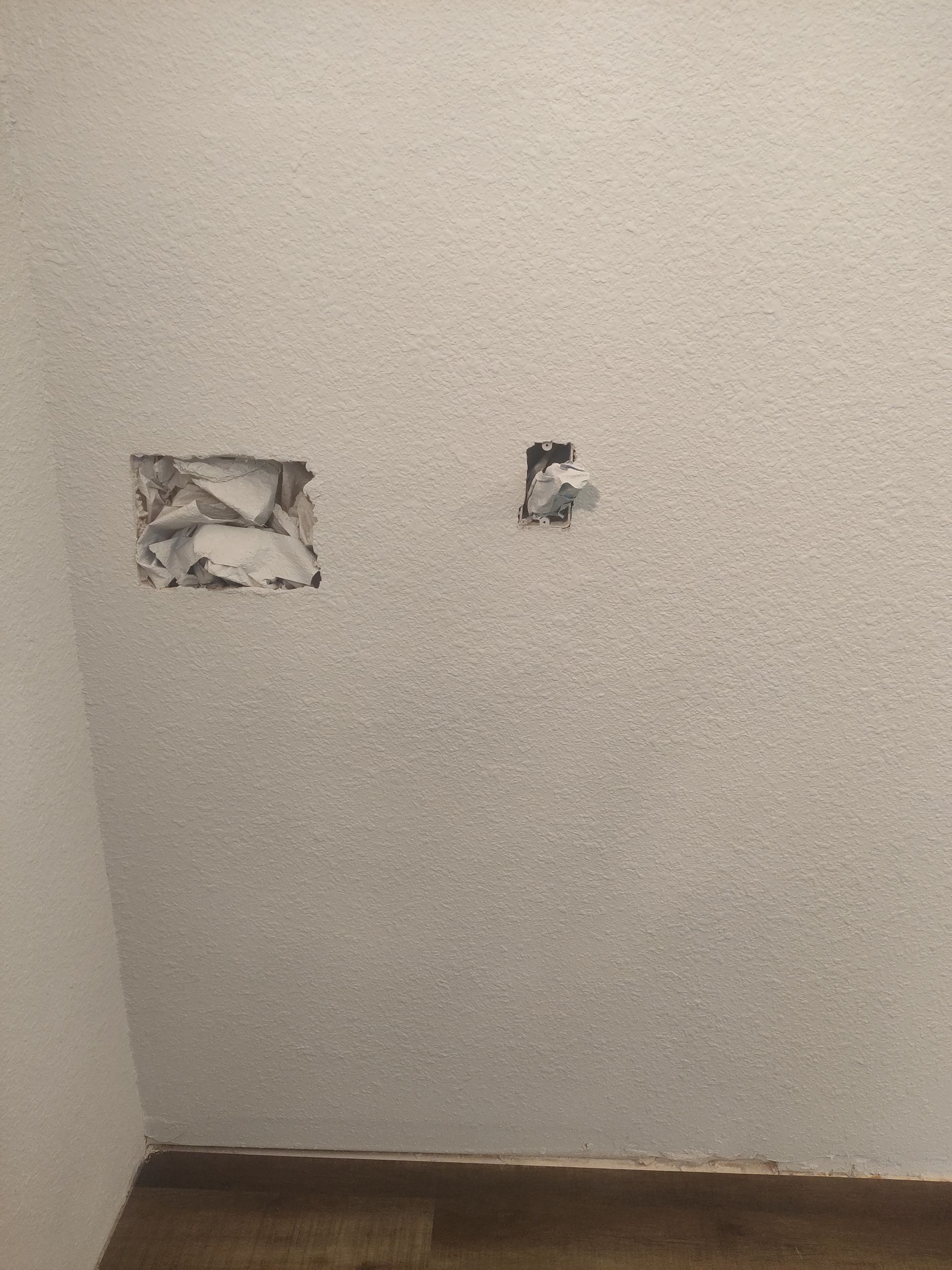 A plain, textured, off-white wall with two rectangular holes cut into the drywall, showing the interior structure.