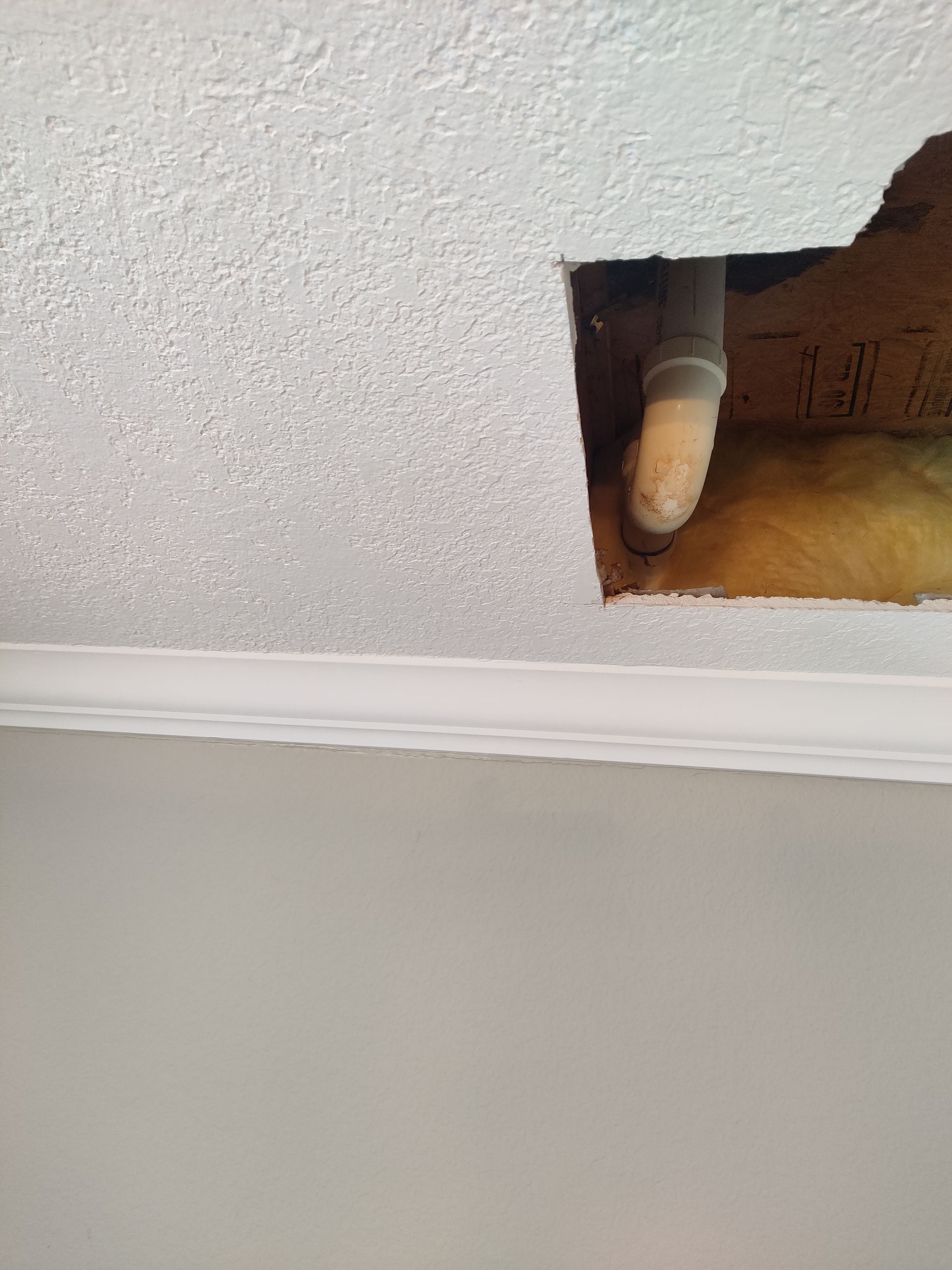 A hole in a textured white ceiling reveals a gray pipe and yellow insulation above, near a crown molding trim.