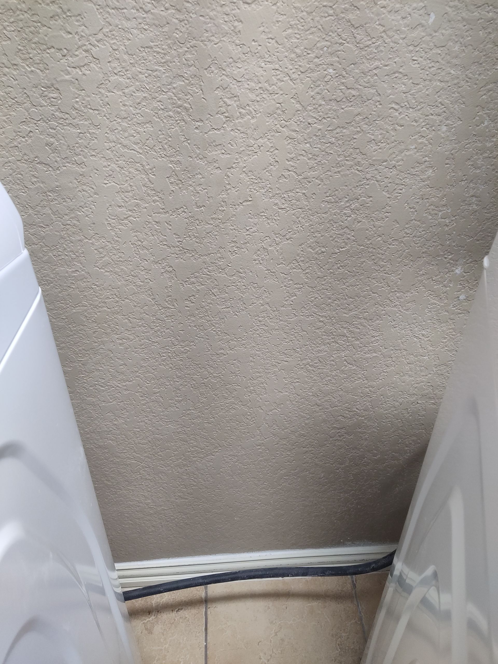 A close-up view of the gap between two white appliances, showing a textured beige wall and a black hose along the floor.