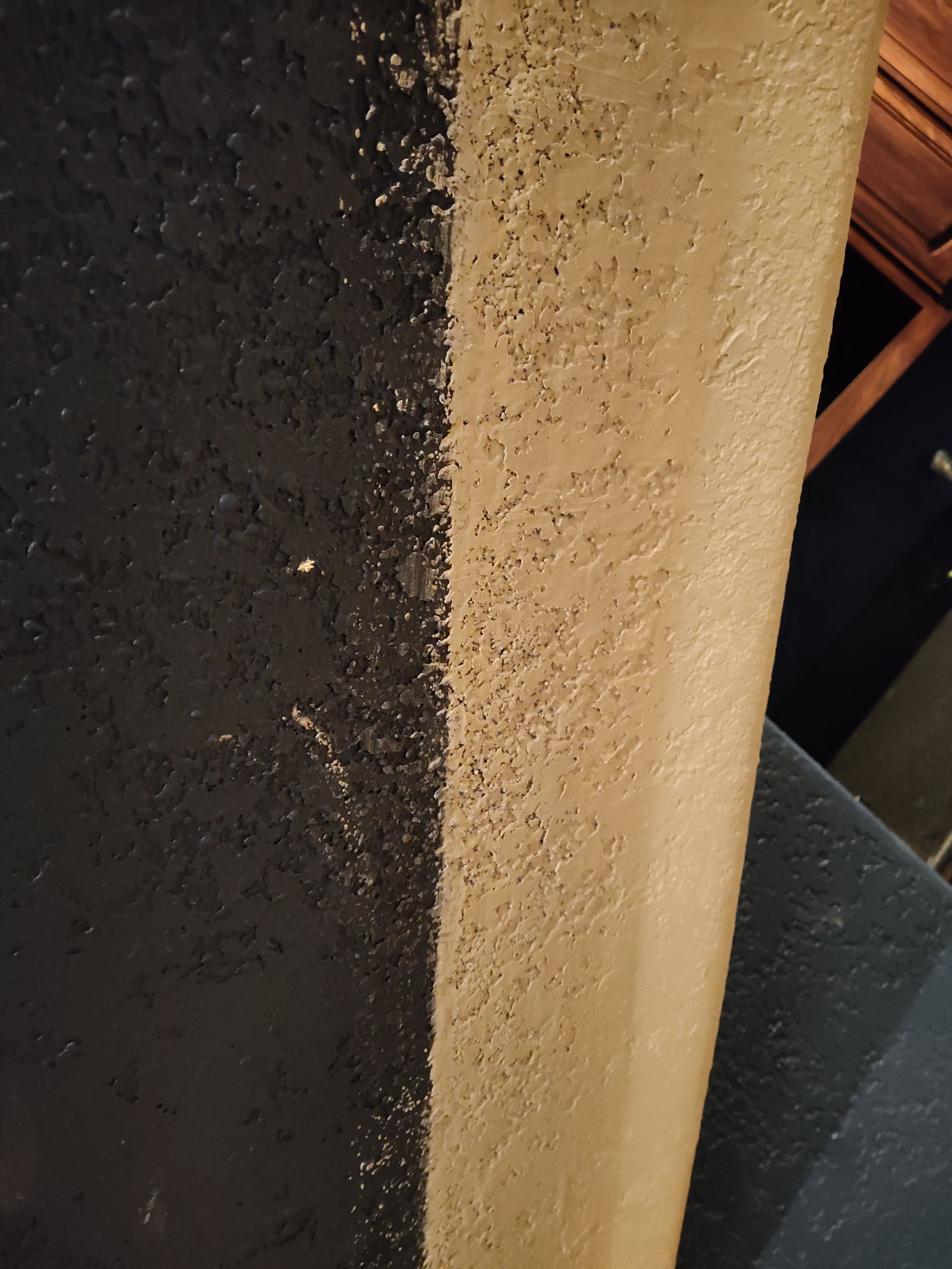 A close-up view shows a vertical transition between a dark, grainy textured surface and a light, tan textured wall.