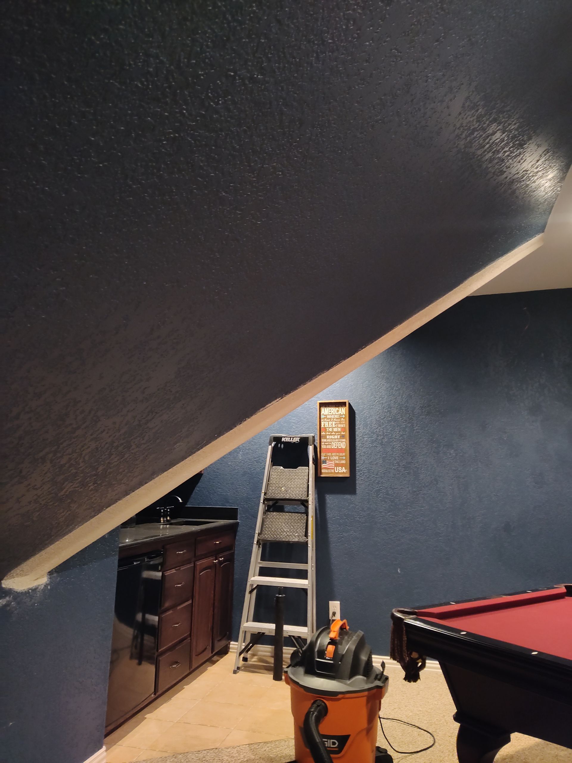 A dark blue room under a sloped ceiling featuring a wet bar, a ladder, a shop vacuum, and a corner of a pool table.