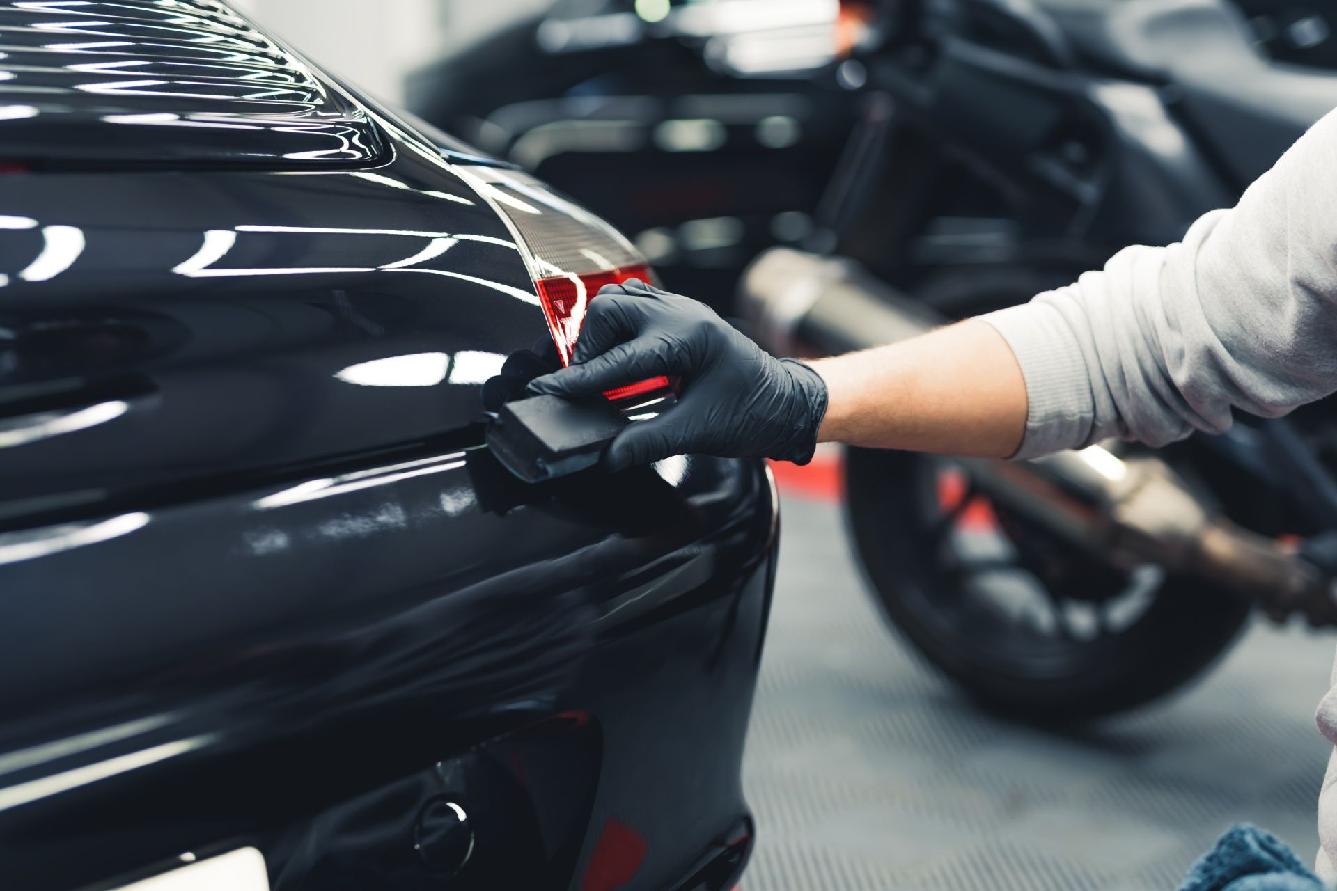 A person is polishing a car in a garage with a motorcycle in the background.