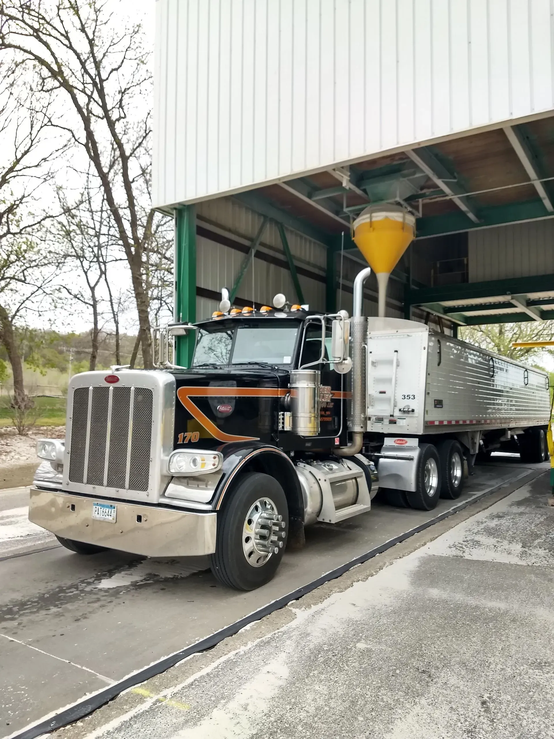 A black Peterbilt semi-truck parked under a loading bay while receiving a load from a yellow funnel.
