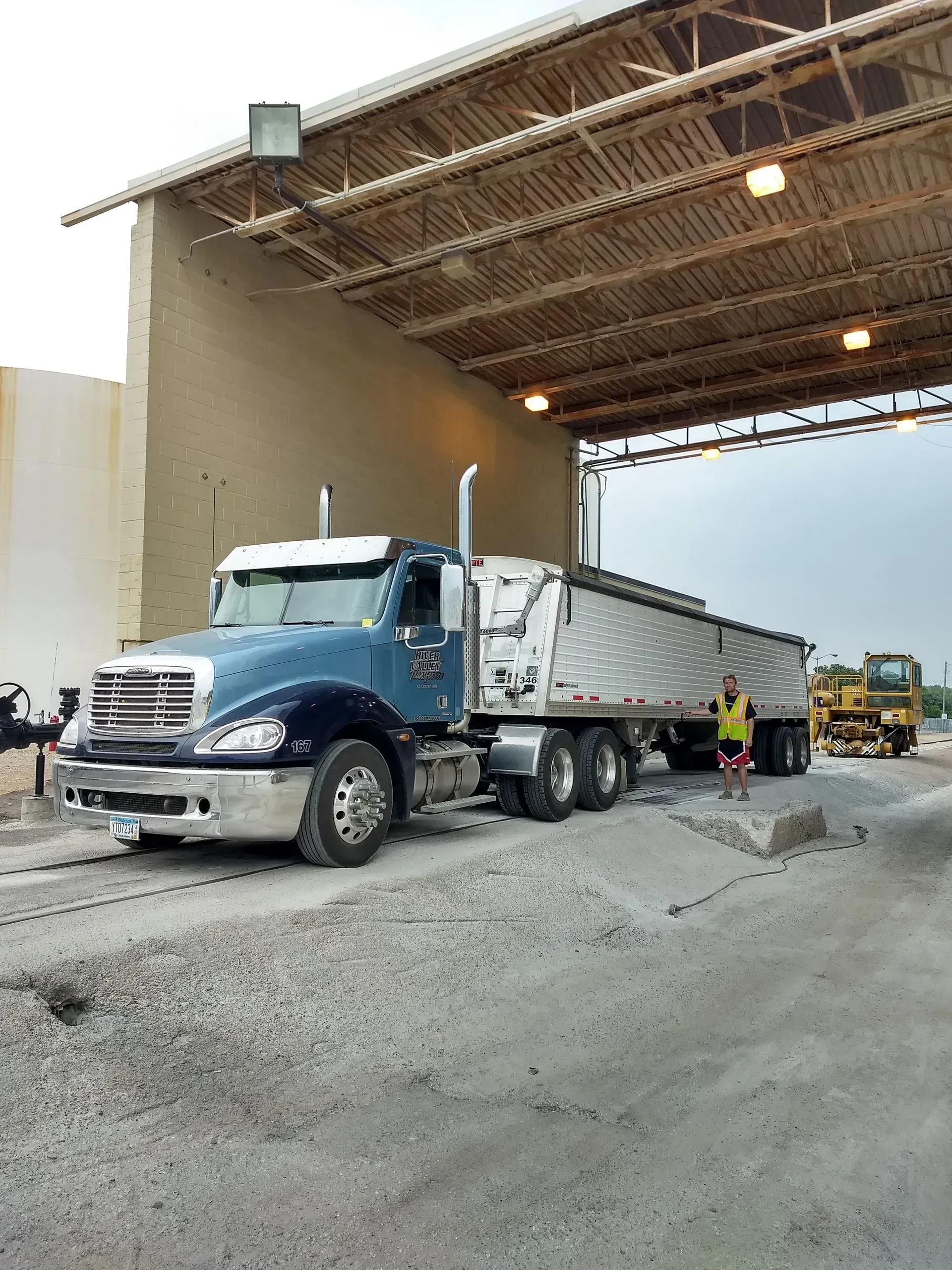 A blue semi-truck parked under an industrial structure, with a worker standing beside the trailer on a gravel lot.