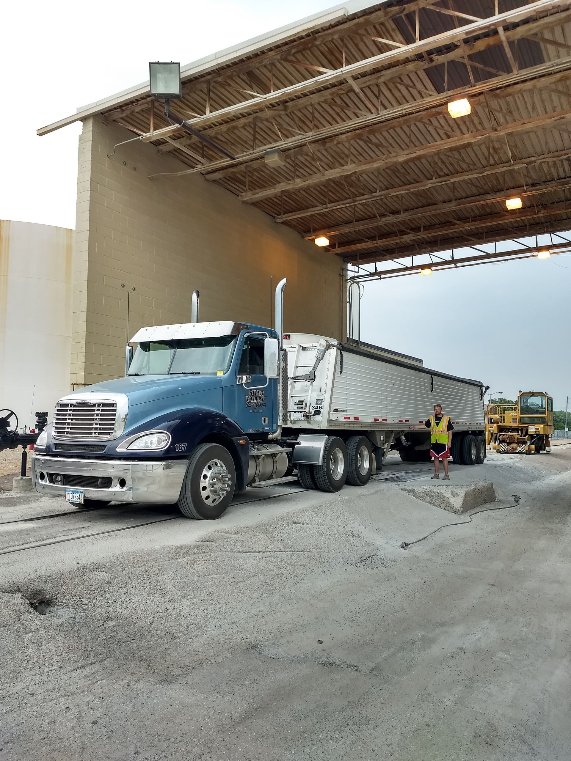A blue semi-truck sits under an industrial canopy on a gravel lot as a person in a safety vest stands nearby.
