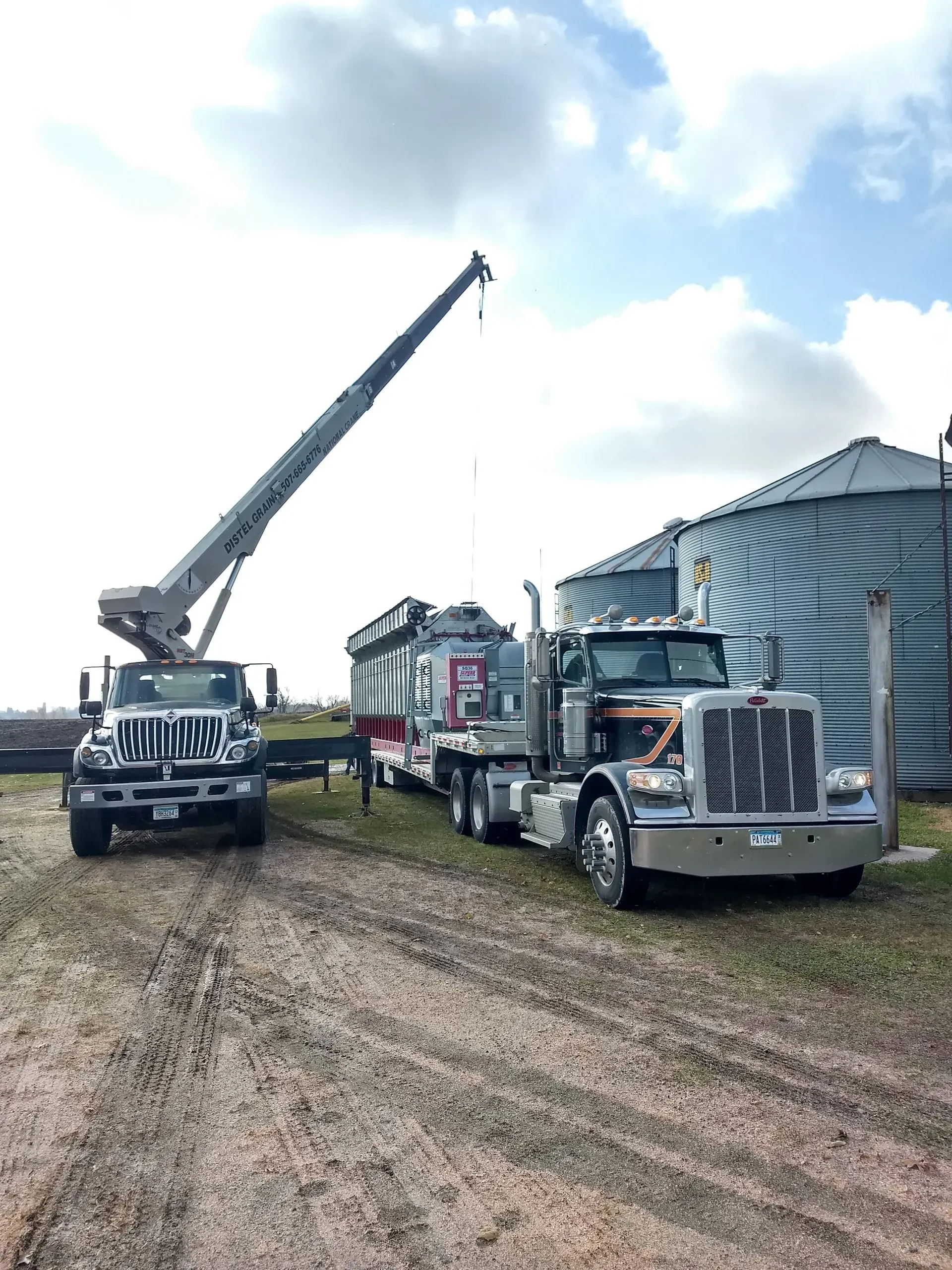 A crane truck and a semi-truck are parked near large grain bins on a dirt lot under a cloudy sky.