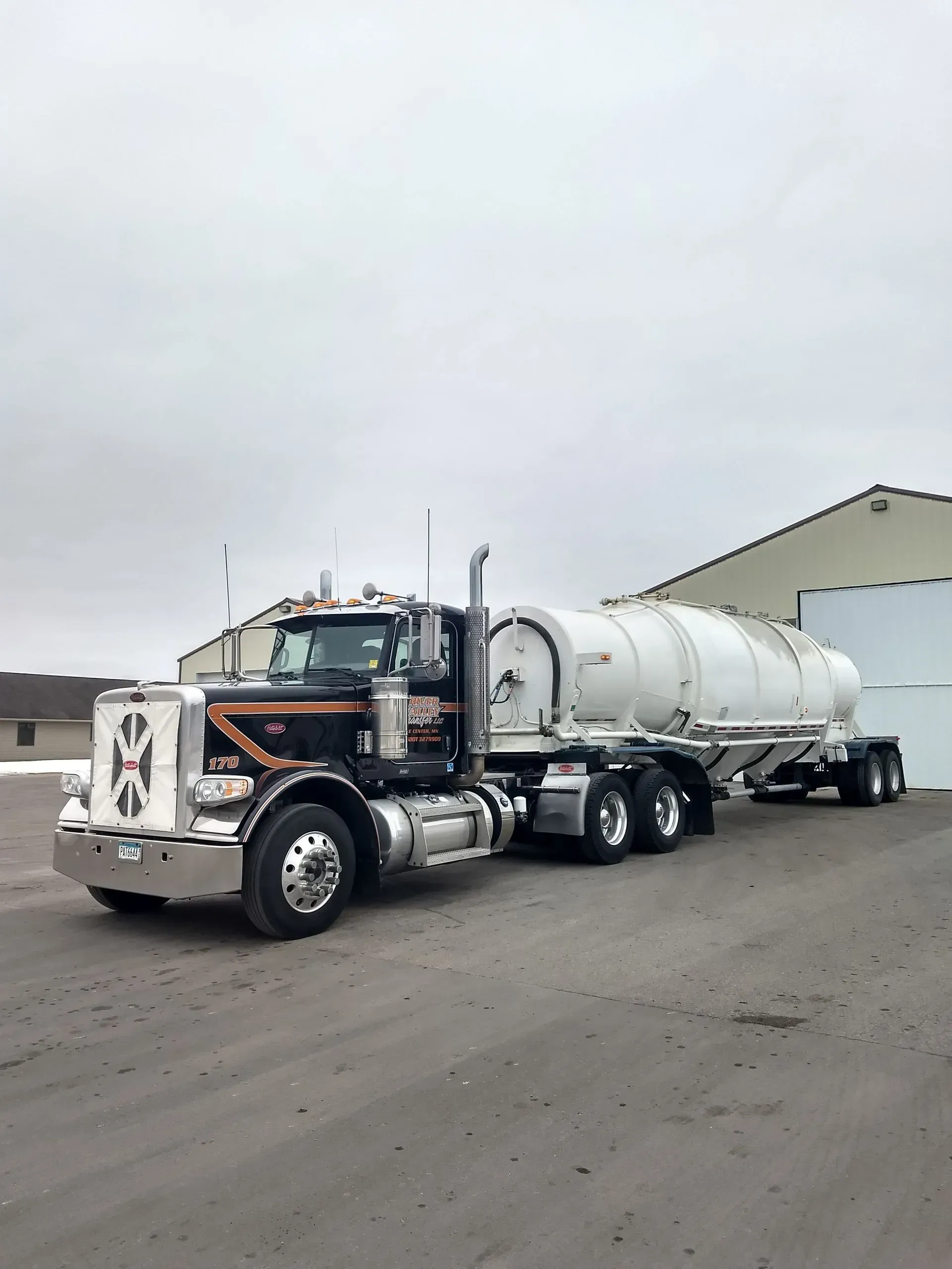 A black semi-truck pulling a large, white cylindrical tank trailer parked on an asphalt lot against an overcast sky.