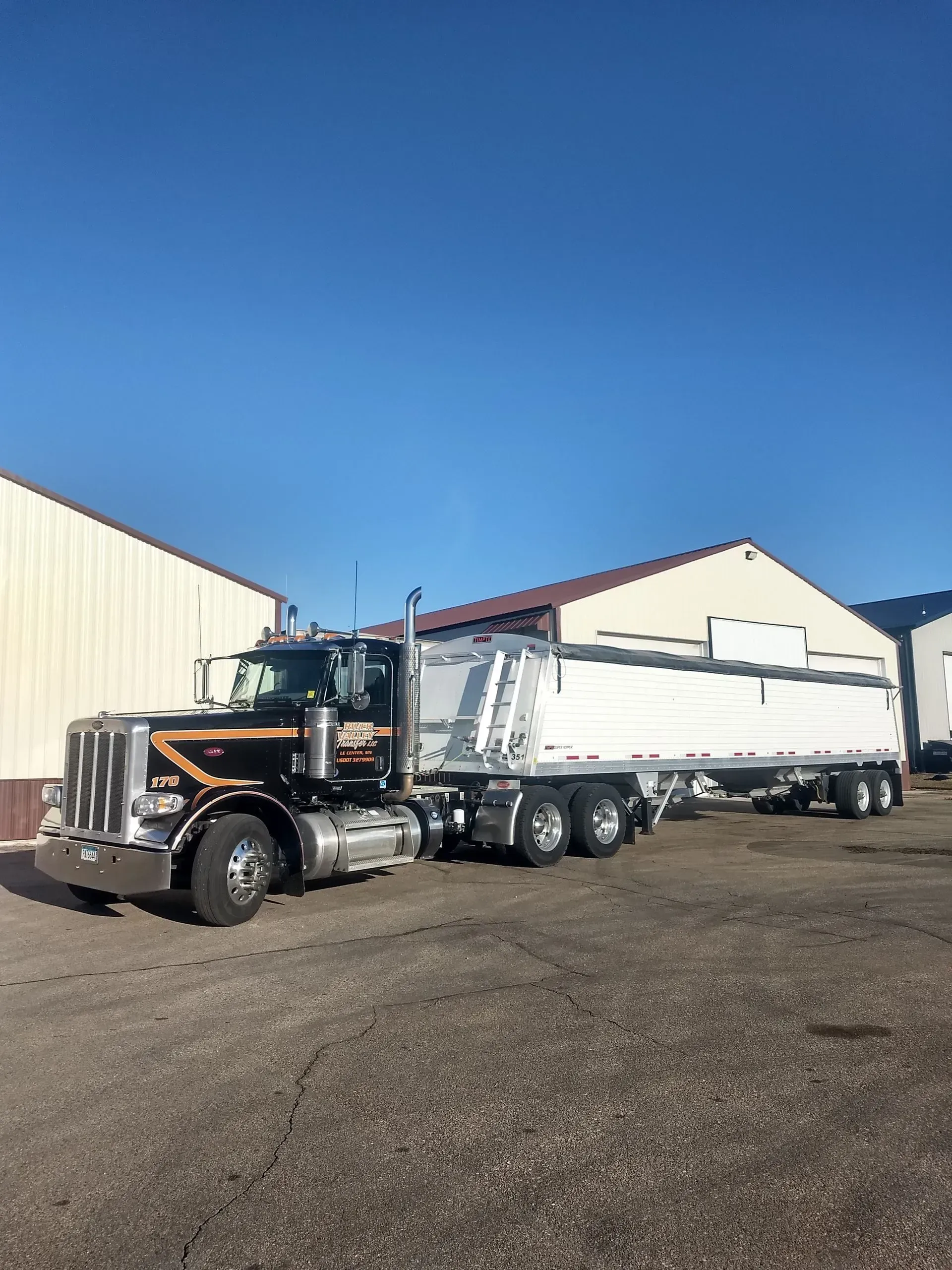 A black semi-truck pulling a long white dump trailer parked in a gravel lot against a clear blue sky.