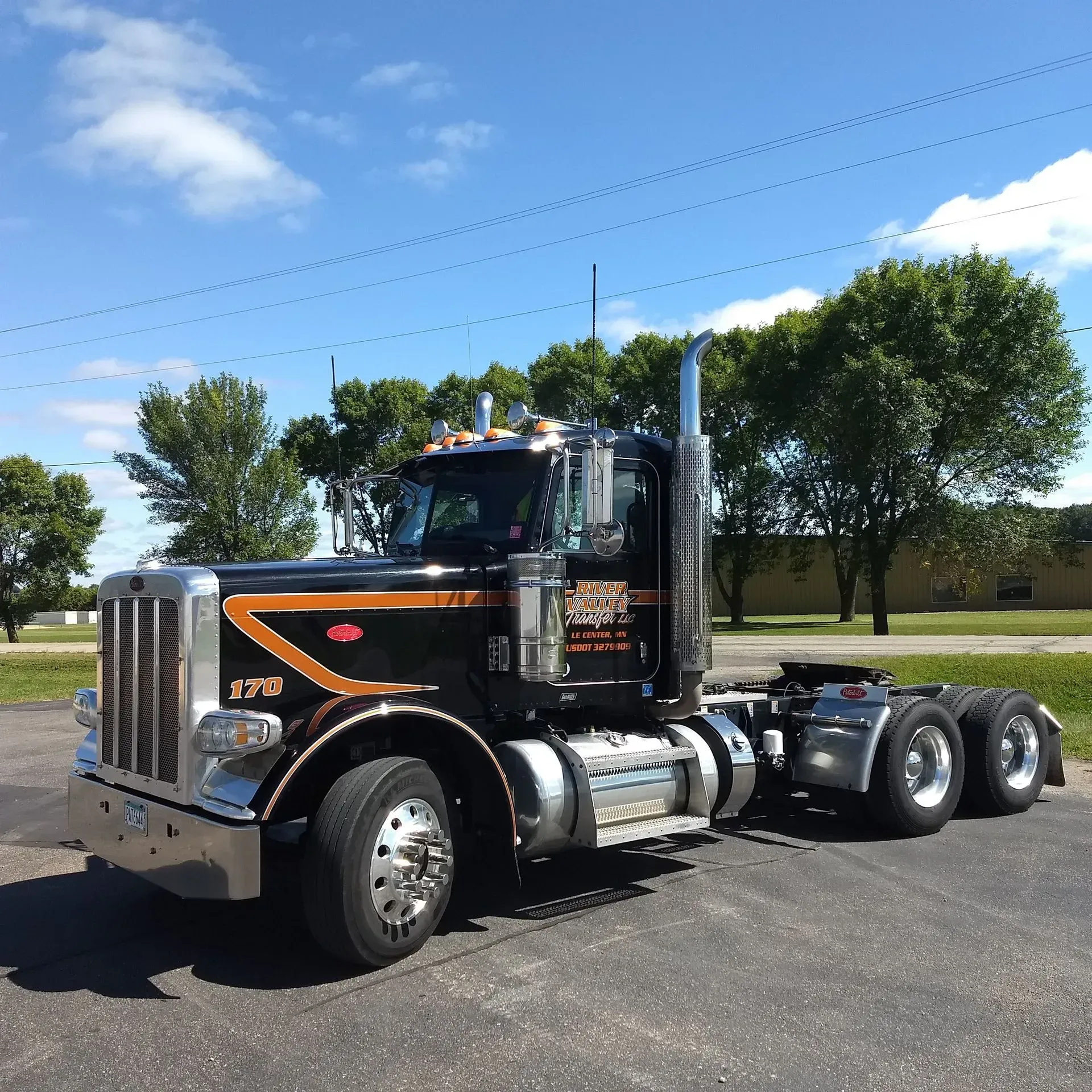 A black Peterbilt semi-truck with orange trim parked on asphalt against a blue sky with trees in the background.