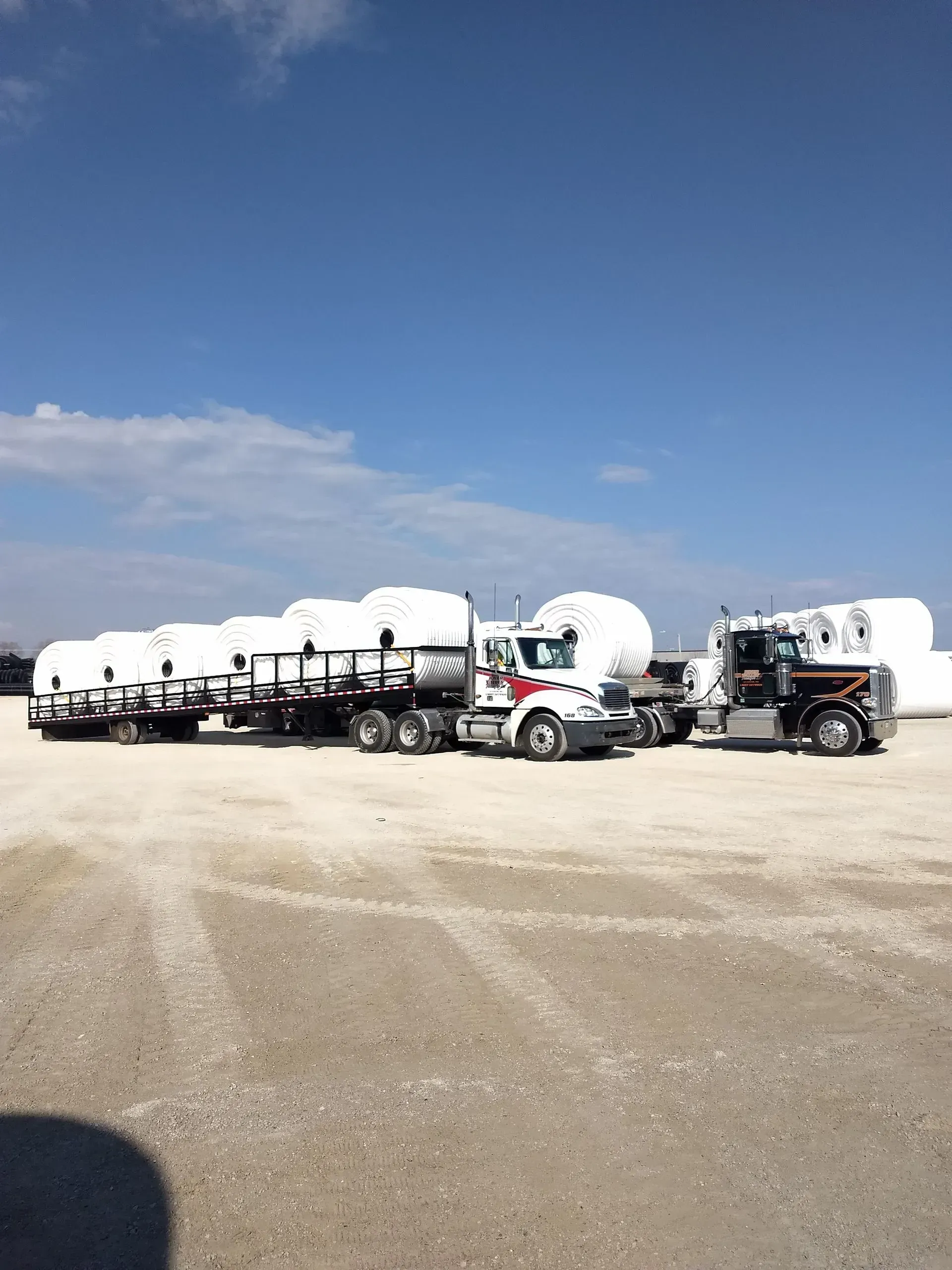 A line of semi-trucks with flatbed trailers carrying large, white cylindrical tanks, parked on a gravel lot under blue sky.
