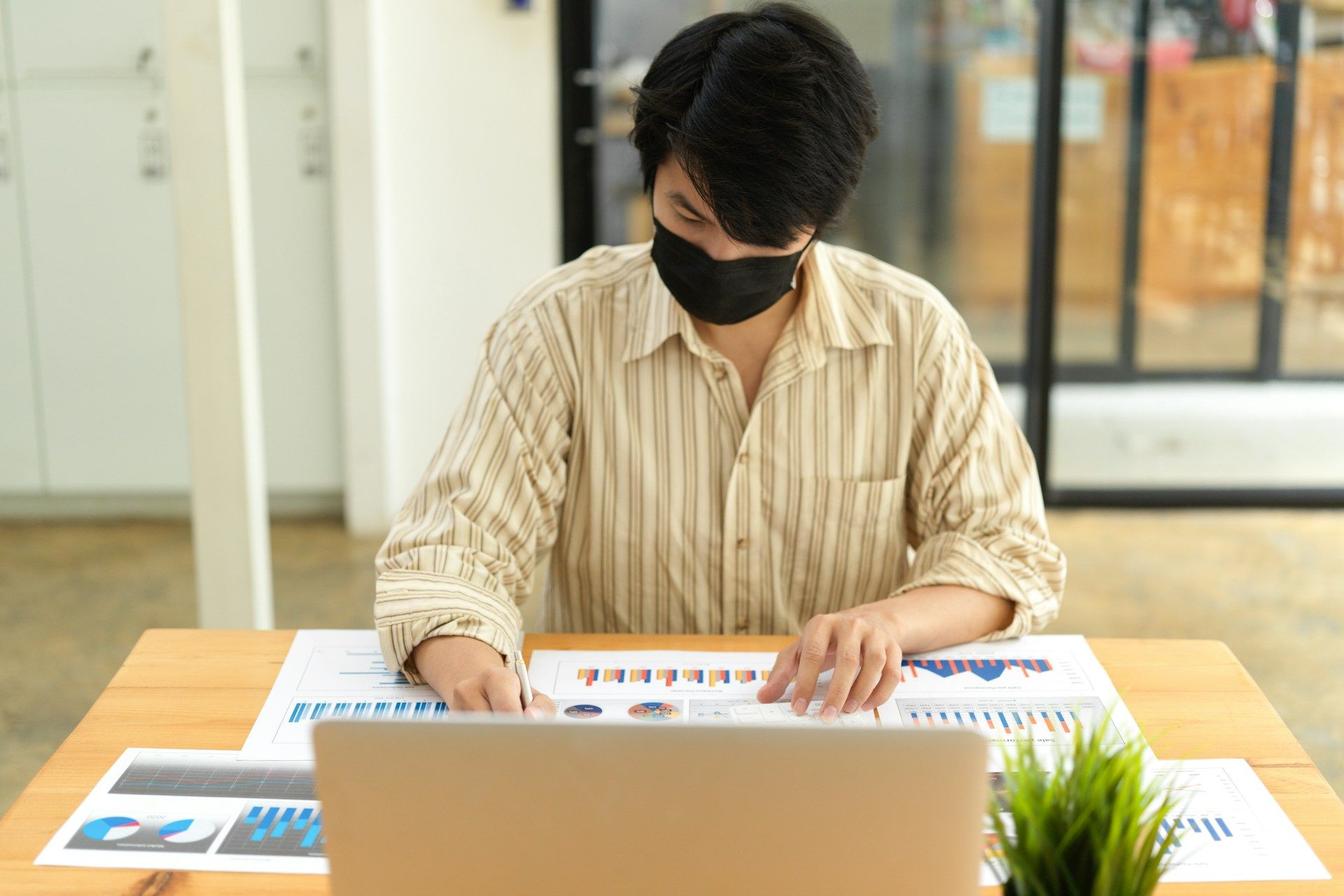 A man wearing a mask is sitting at a desk using a laptop computer.