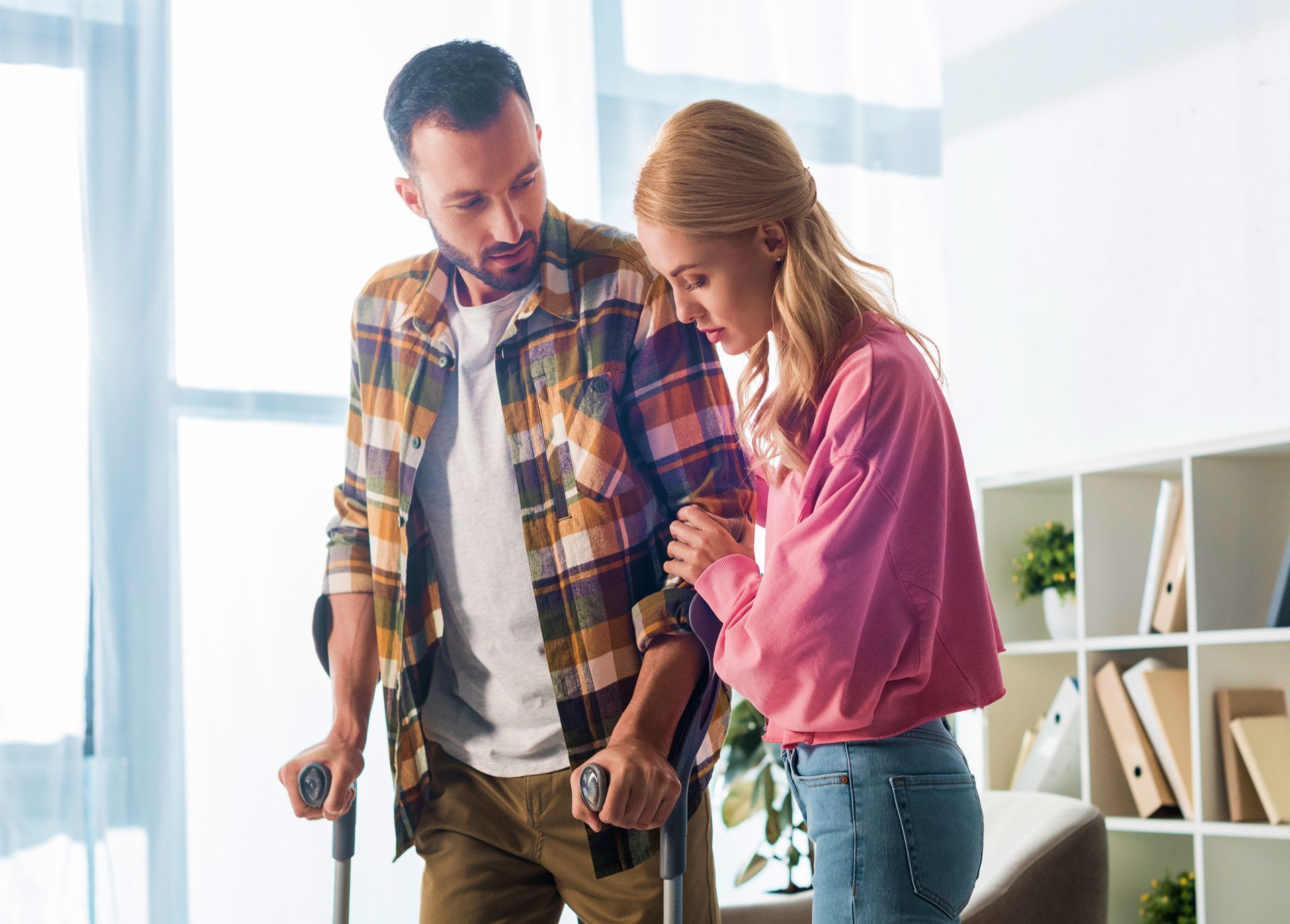 A woman is helping a man with crutches walk in a living room.