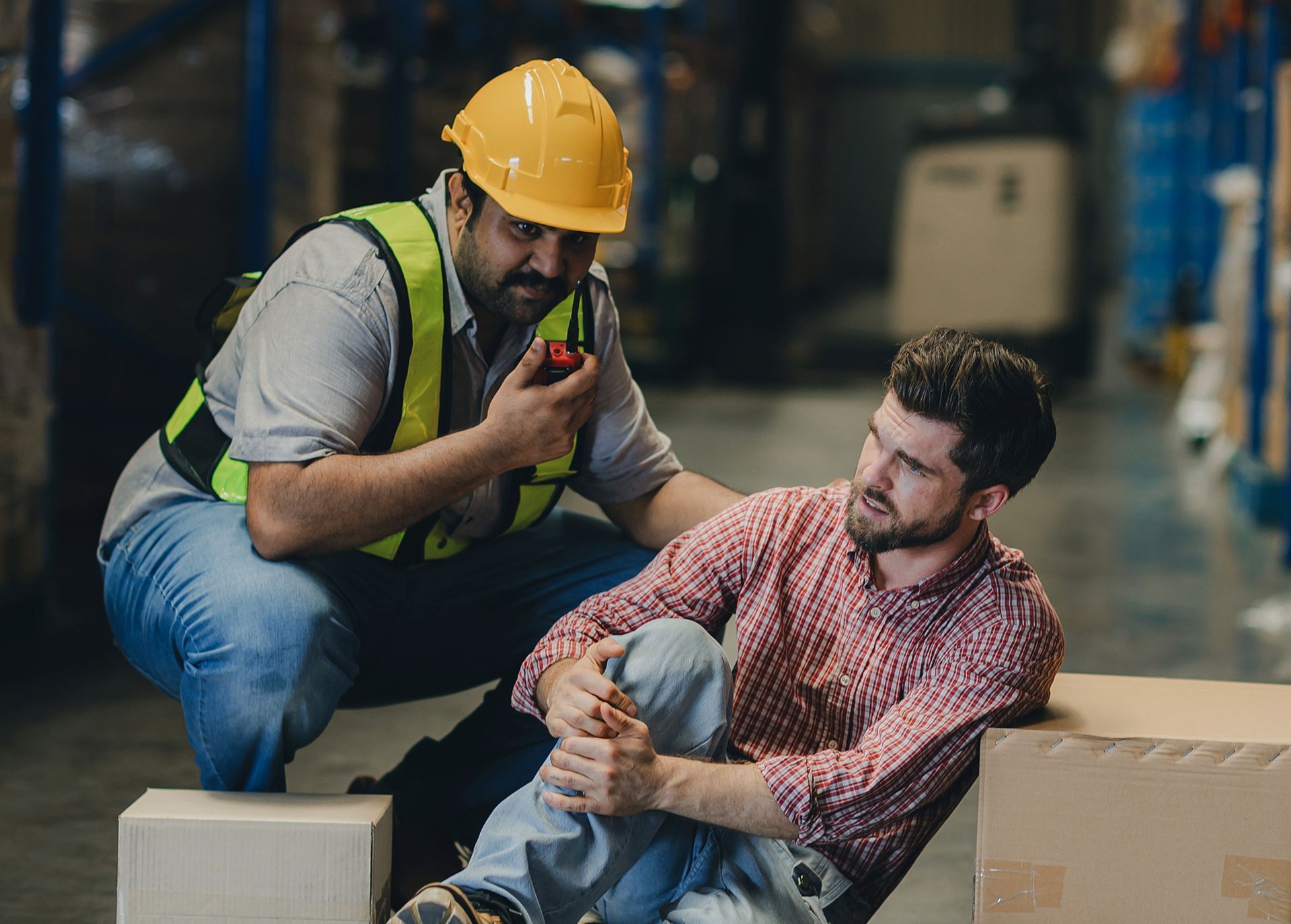 A man in a hard hat is helping another man who has fallen in a warehouse.