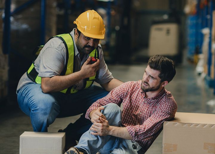 A man in a hard hat is helping another man who has fallen in a warehouse.