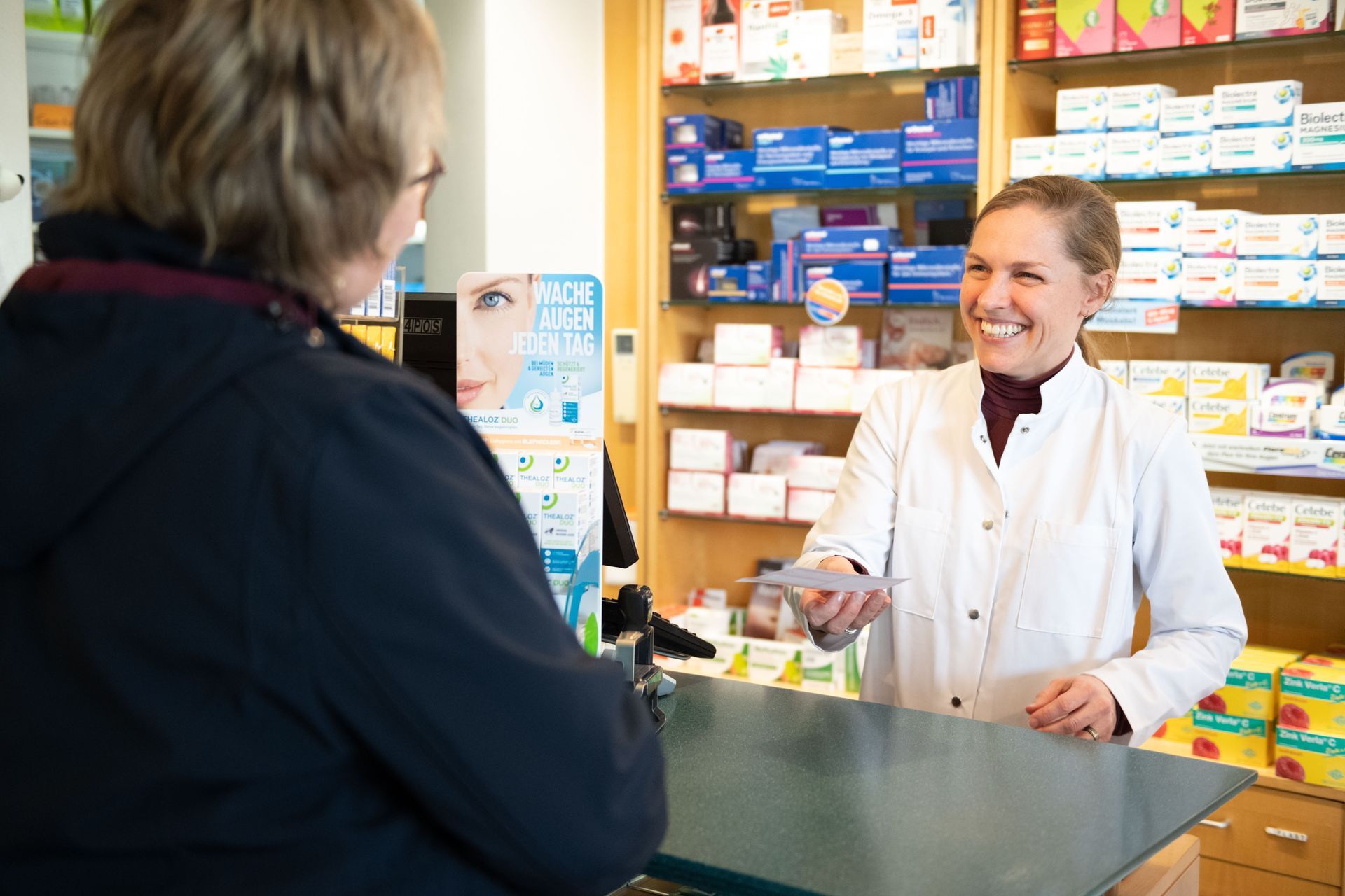 Eine Frau steht an einem Tresen in einer Apotheke und spricht mit einem Kunden.
