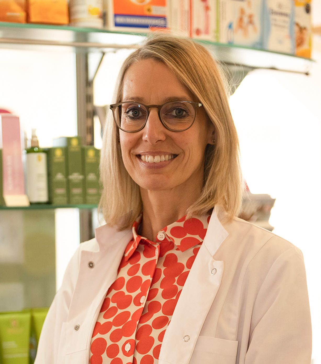 Eine Frau mit Brille und Laborkittel lächelt in einer Apotheke