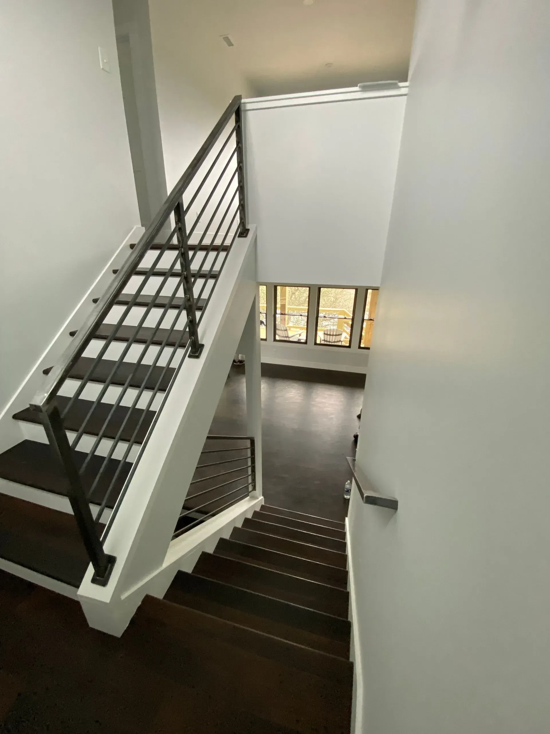 Staircase with dark wood steps and a metal railing, leading down to a living area with windows. The walls are painted white.