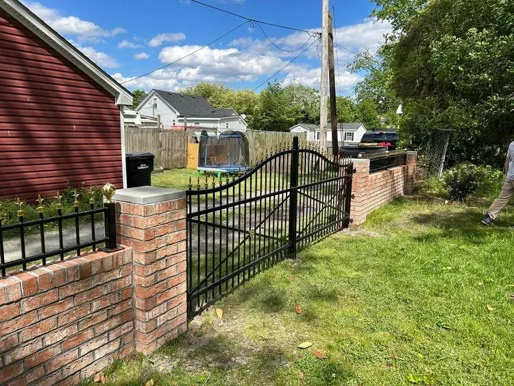 A black wrought iron gate in a brick wall leads into a grassy yard with a trampoline, with a red building to the left.